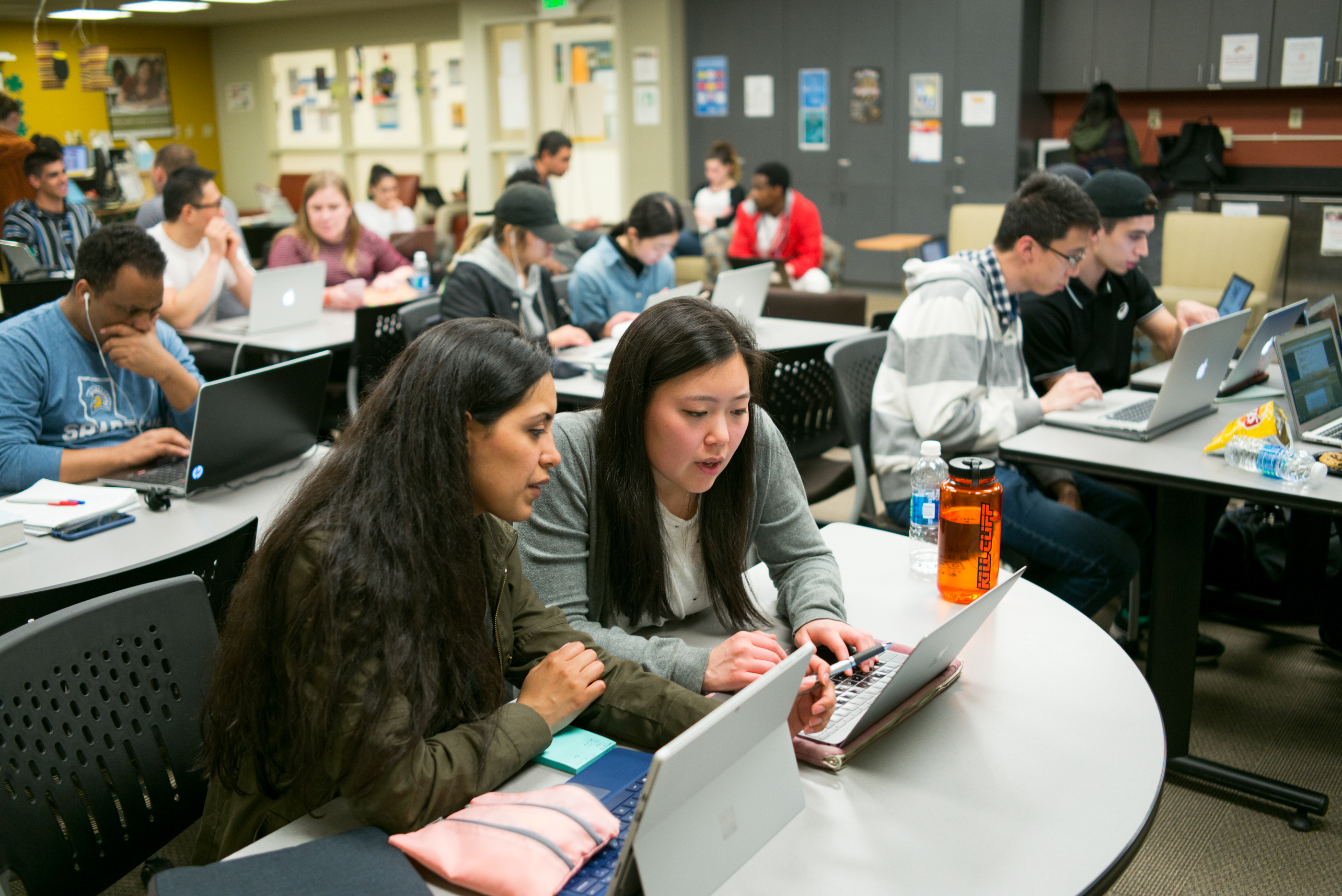 Two female students sit in a large classroom looking at information on their computer screens. 