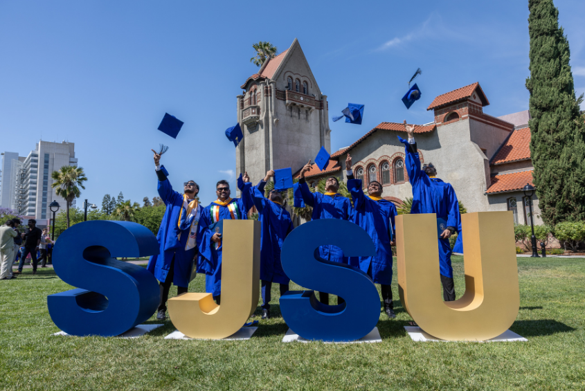 Students tossing their caps in the air while standing behind SJSU lettering