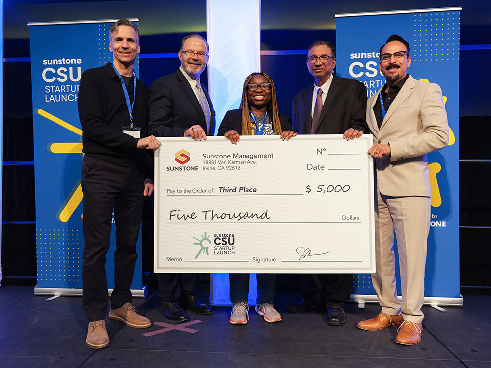 A female, college-age startup founder poses onstage with two sponsors, two hosts, and a giant check for $5,000