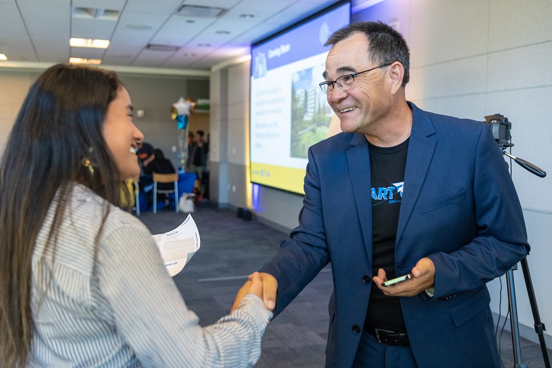 A woman in a striped blouse with long, dark hair shakes hands with a smiling man with short, salt-and-pepper hair.  He wears a black t-shirt with the SpartUp logo under a blue suit. 