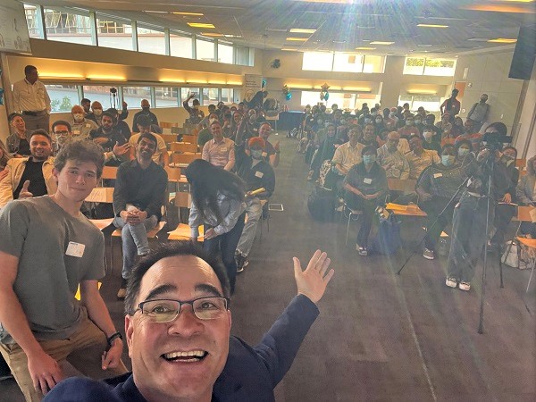 A dark haired man dressed in a sport coat and tshirt, laughing, takes a selfie with an amber-toned roomful of people behind him. 