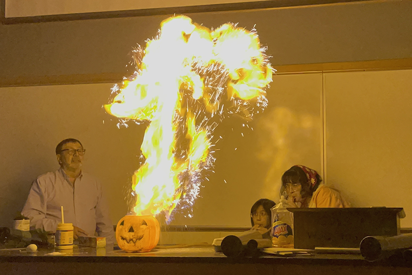 Photograph of Alejandro Garcia behind a fireball of burning corn starch, emerging from a jack-o-lantern.