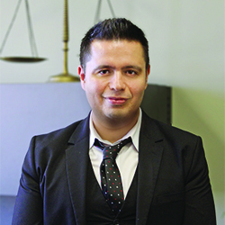 Hispanic male with short, black hair, wearing a black suit with a white, button-down shirt and a white spotted, black tie, Smiling in front of brass-colored justice scales.  