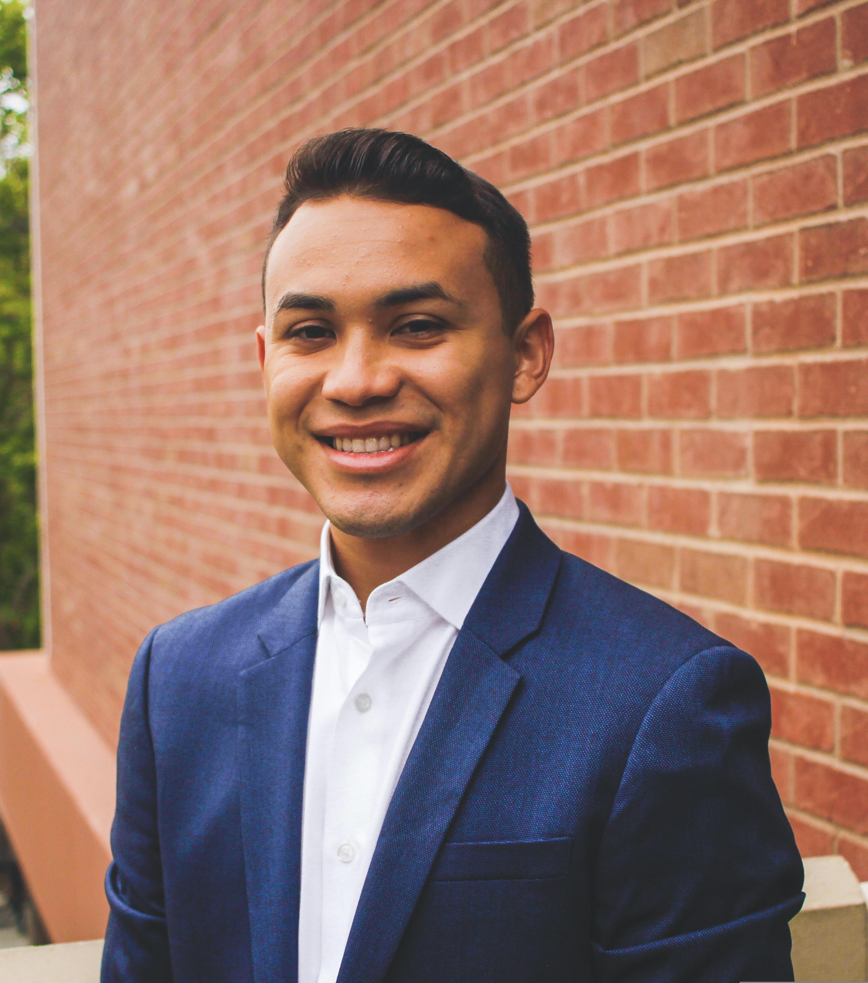 Hispanic male with short, black hair, wearing a blue suit jacket and white button down, smiling in front of a red brick wall.