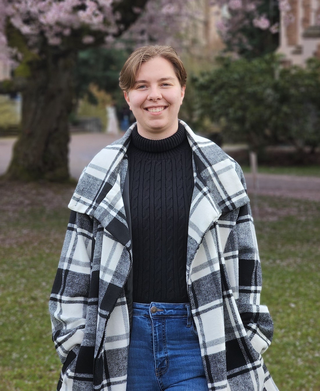 White woman, in her twenties, with short, dirty blonde hair, wearing a checkered coat and black sweater smiling in front of pink cherry blossoms.