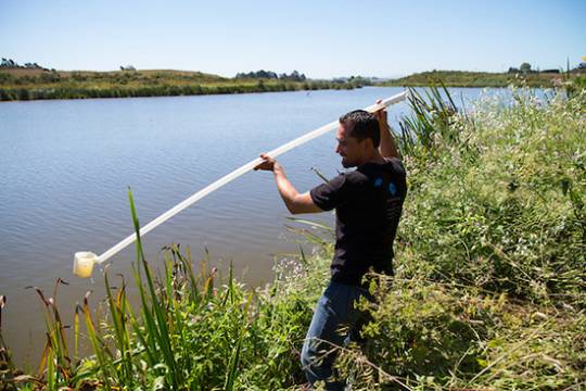 person draws sample water from a sizeable river with a long pole