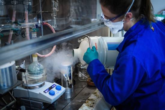 person in lab coat and gear pours big vessel of steaming substance into smaller container under fume hood