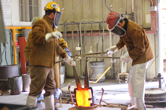 people in foundry gear hold large glowing crucible with metal tongs in foundry with equipment and gear all around 