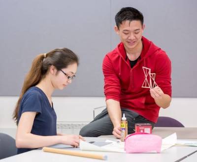 students work together.  One sits at table, one sits on table.  they work with wood pieces and glue.