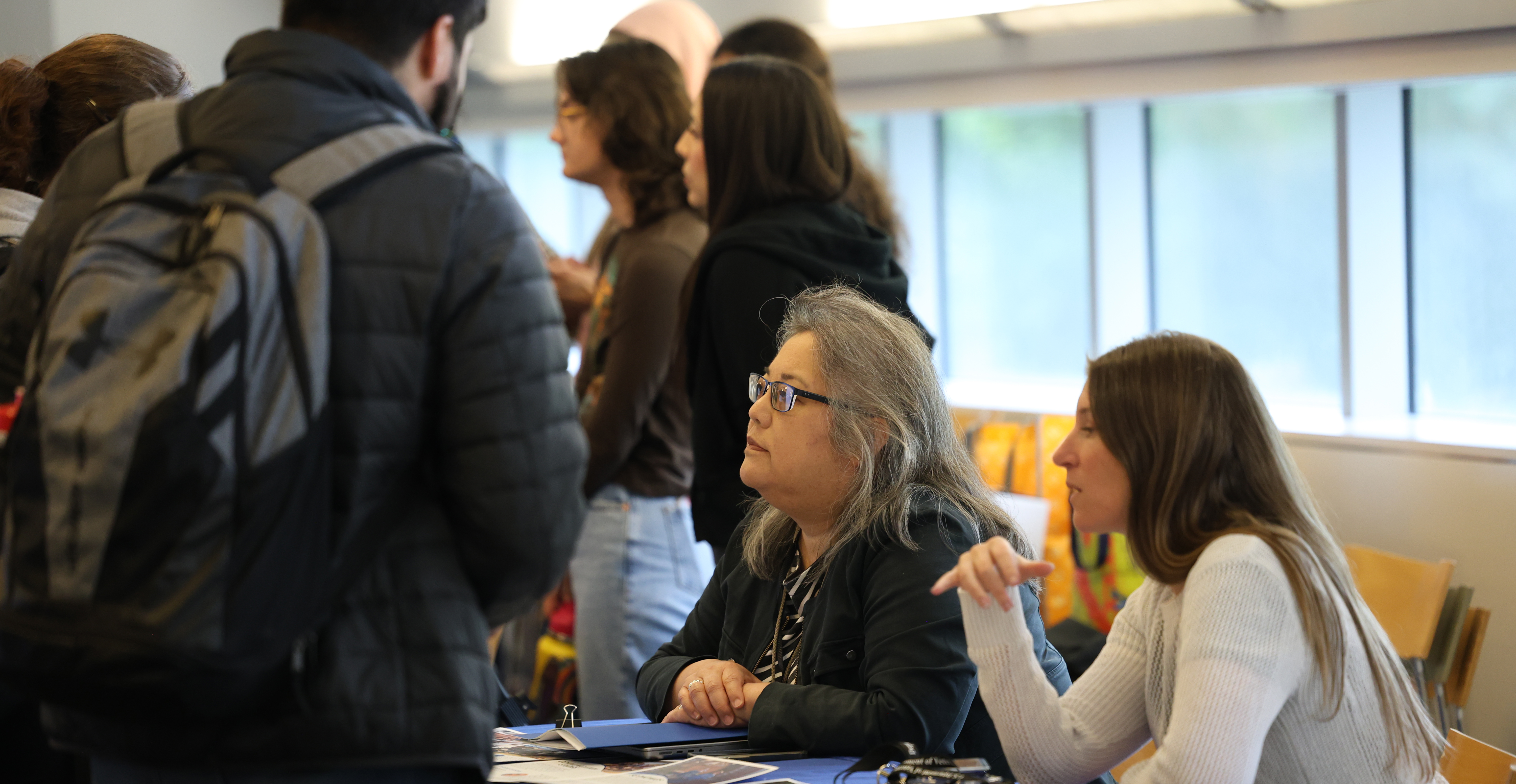 A professor and staff member or student assistant sit at table full of materials, talking to students.  More tablers and students are in back.