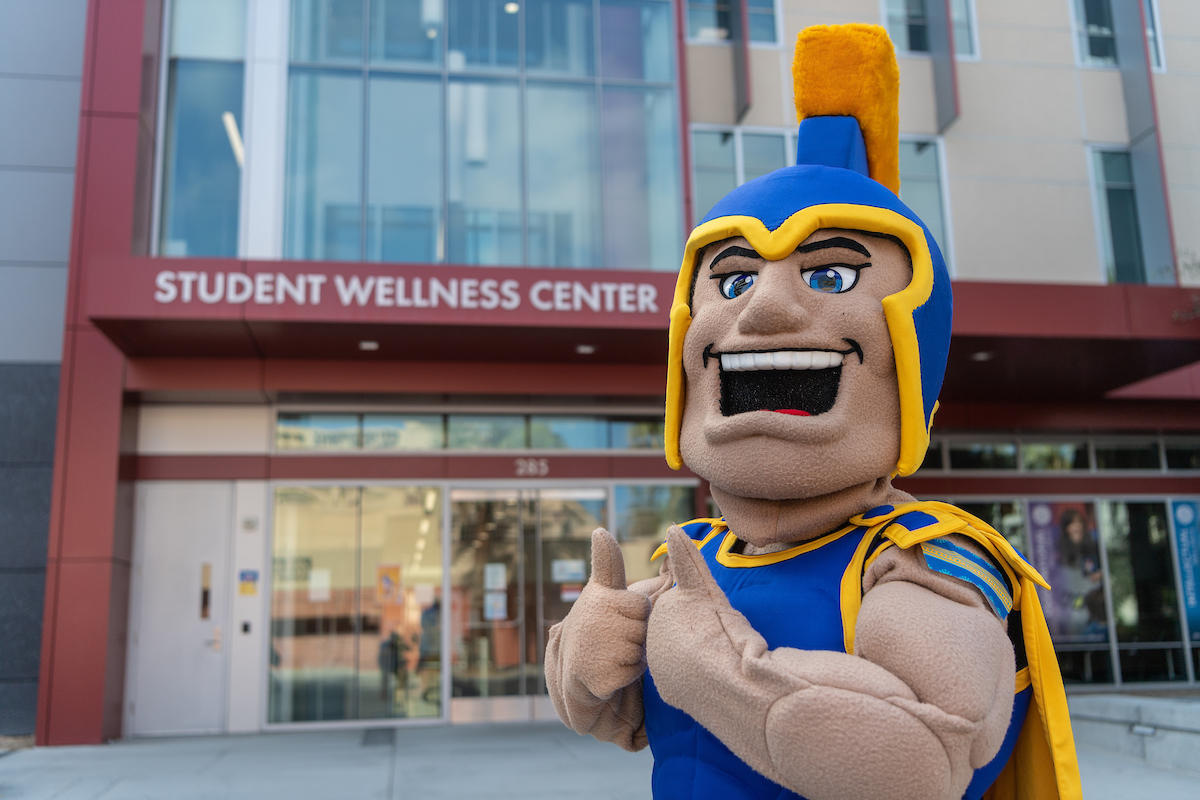 Sammy with their thumbs up standing in front of the Student Wellness Center